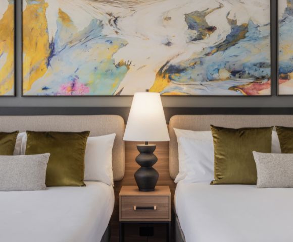 Two neatly made beds with white linens and green pillows flank a wooden nightstand with a black lamp. Abstract art with blue and yellow splashes hangs above.