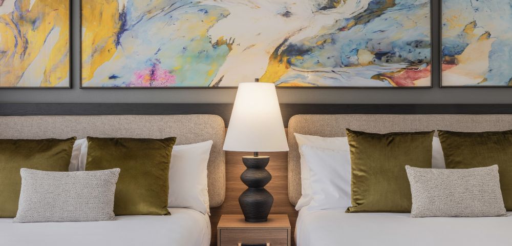 Two neatly made beds with white linens and green pillows flank a wooden nightstand with a black lamp. Abstract art with blue and yellow splashes hangs above.