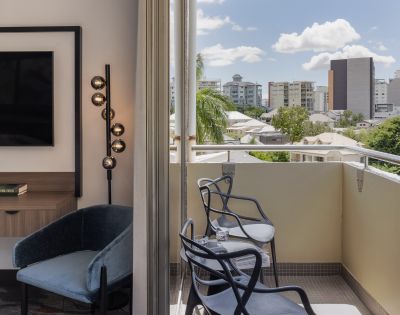 Modern room with a balcony overlooking a cityscape. Chairs and table on the balcony; city buildings and palm trees visible under a blue sky with clouds.