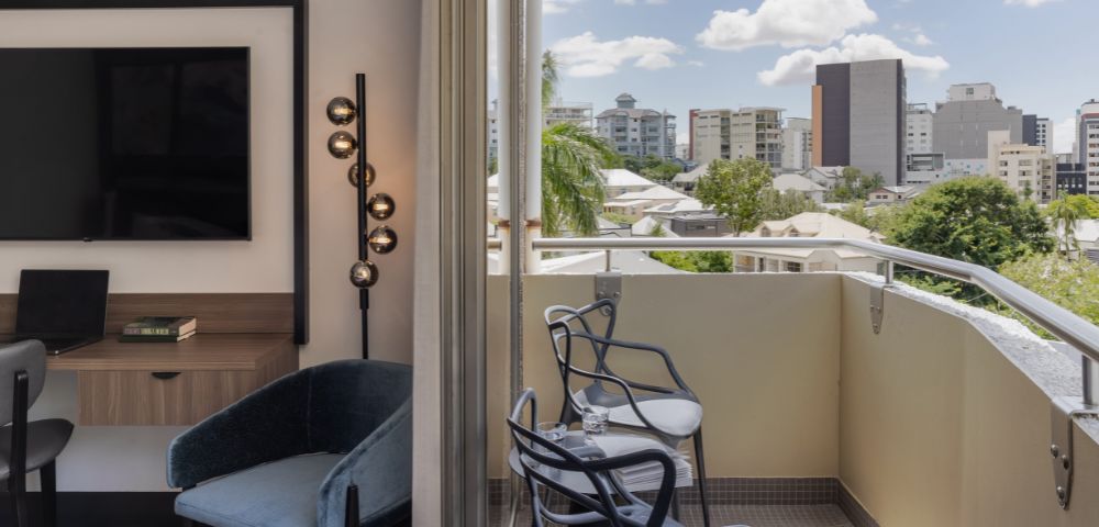 Modern room with a balcony overlooking a cityscape. Chairs and table on the balcony; city buildings and palm trees visible under a blue sky with clouds.