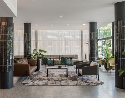Modern lobby with sleek black columns and elegant seating. Neutral-toned sofas and armchairs surround a decorative rug. Large windows bring in natural light.