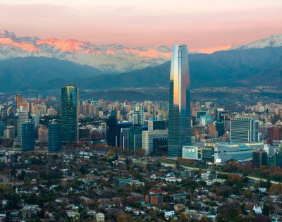 Panoramic view of Santiago, Chile's skyline at sunrise, featuring modern skyscrapers, green spaces, and the Andes mountains bathed in pink light.