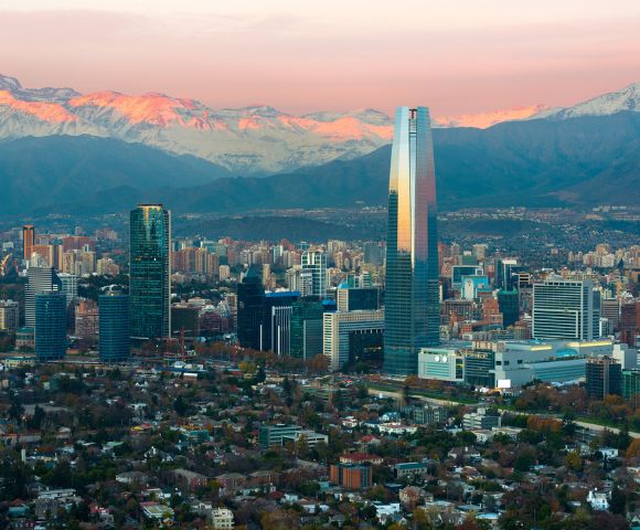 Panoramic view of Santiago, Chile's skyline at sunrise, featuring modern skyscrapers, green spaces, and the Andes mountains bathed in pink light.
