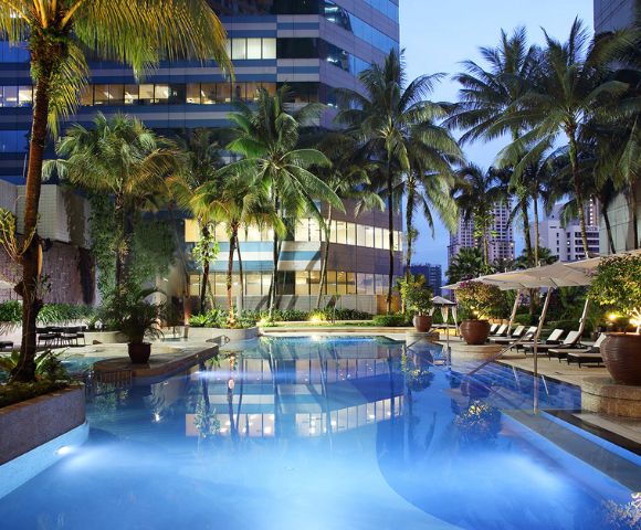 Luxurious hotel pool at twilight surrounded by palm trees and modern high-rise buildings. Lounge chairs and umbrellas create a serene, inviting atmosphere.