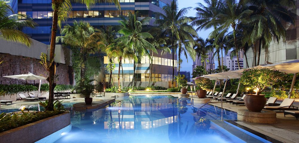 Luxurious hotel pool at twilight surrounded by palm trees and modern high-rise buildings. Lounge chairs and umbrellas create a serene, inviting atmosphere.