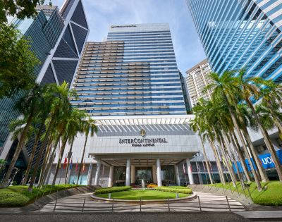 Front view of the InterContinental Hotel Kuala Lumpur, featuring a modern glass facade, flanked by lush palm trees under a clear blue sky.