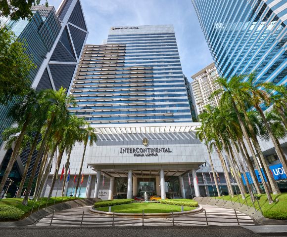 Front view of the InterContinental Hotel Kuala Lumpur, featuring a modern glass facade, flanked by lush palm trees under a clear blue sky.