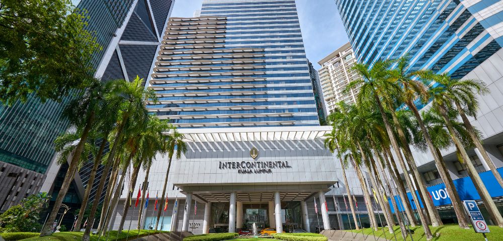 Front view of the InterContinental Hotel Kuala Lumpur, featuring a modern glass facade, flanked by lush palm trees under a clear blue sky.