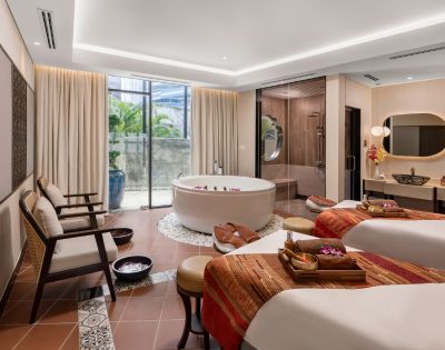 Elegant spa room with two massage tables draped in warm, patterned blankets. A round bathtub sits beside them. Soft lighting creates a tranquil ambiance.