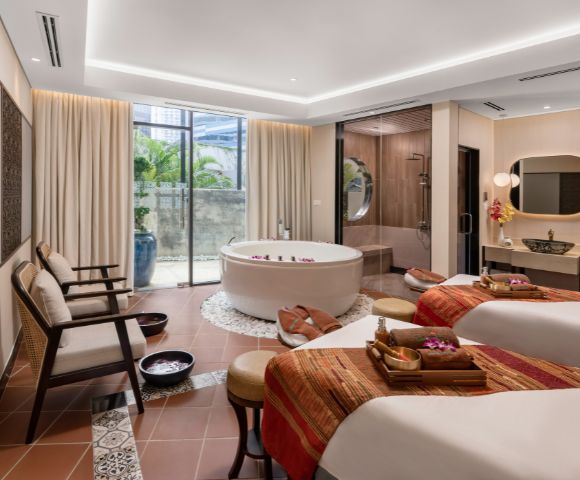 Elegant spa room with two massage tables draped in warm, patterned blankets. A round bathtub sits beside them. Soft lighting creates a tranquil ambiance.