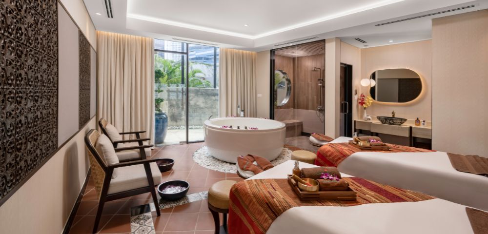 Elegant spa room with two massage tables draped in warm, patterned blankets. A round bathtub sits beside them. Soft lighting creates a tranquil ambiance.
