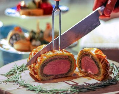 A knife slices into a golden brown Beef Wellington on a wooden board garnished with rosemary. A background tea tray and blurred food suggest elegance.