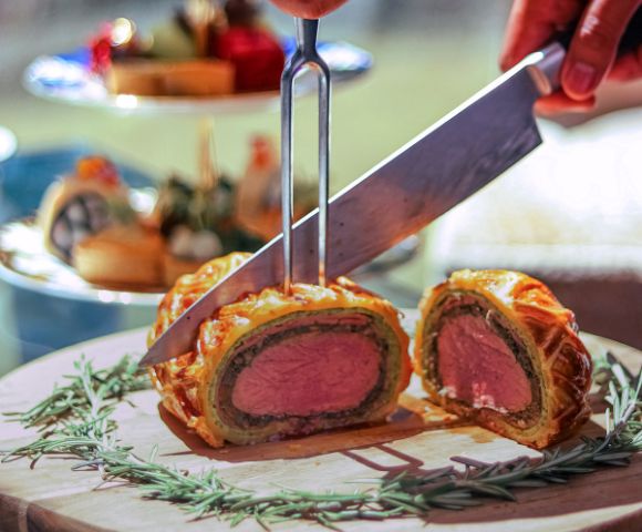 A knife slices into a golden brown Beef Wellington on a wooden board garnished with rosemary. A background tea tray and blurred food suggest elegance.