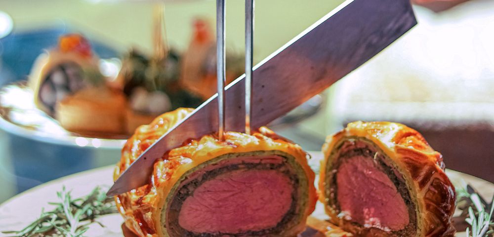 A knife slices into a golden brown Beef Wellington on a wooden board garnished with rosemary. A background tea tray and blurred food suggest elegance.