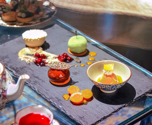 Elegant dessert assortment on a slate platter includes a green mousse sphere, a caramel tart, and decorative cherry garnishes, exuding a luxurious, gourmet vibe.