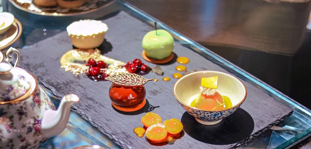 Elegant dessert assortment on a slate platter includes a green mousse sphere, a caramel tart, and decorative cherry garnishes, exuding a luxurious, gourmet vibe.