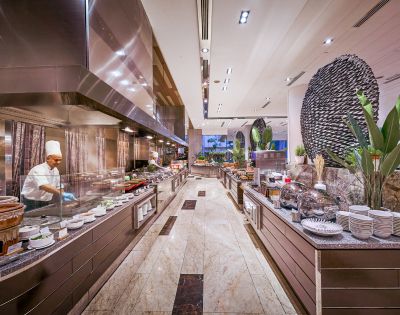 Luxurious buffet restaurant with a chef in a white uniform cooking on the left. Sleek counters, various dishes, tall plants create an elegant ambiance.