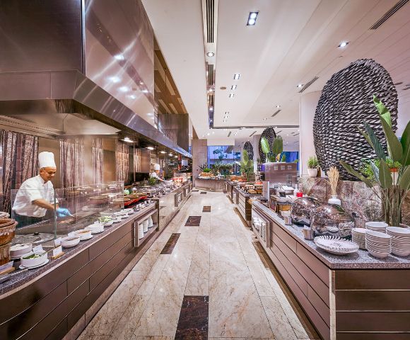 Luxurious buffet restaurant with a chef in a white uniform cooking on the left. Sleek counters, various dishes, tall plants create an elegant ambiance.