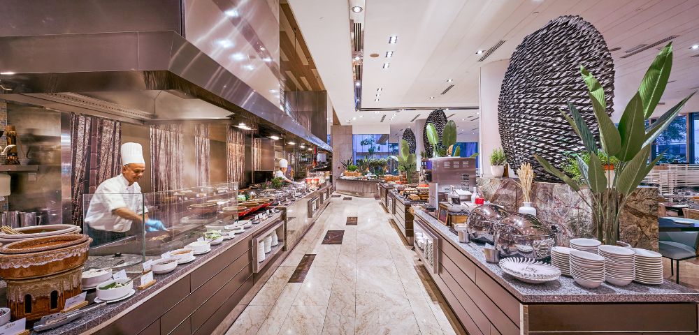 Luxurious buffet restaurant with a chef in a white uniform cooking on the left. Sleek counters, various dishes, tall plants create an elegant ambiance.