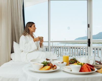 A woman in a robe sits on a bed with breakfast, holding a mug. She gazes out a window at a serene ocean view, creating a calm, relaxed atmosphere.