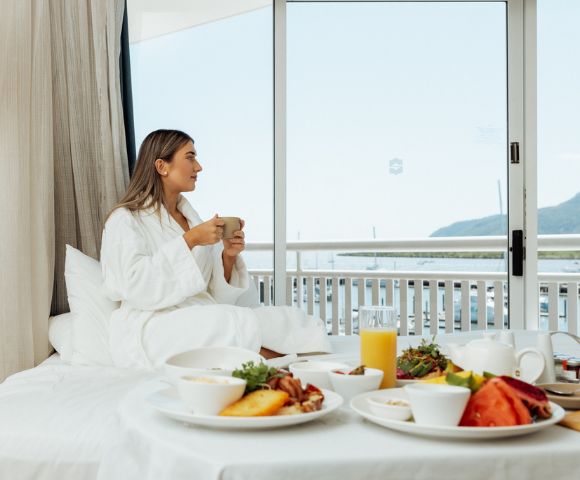 A woman in a robe sits on a bed with breakfast, holding a mug. She gazes out a window at a serene ocean view, creating a calm, relaxed atmosphere.