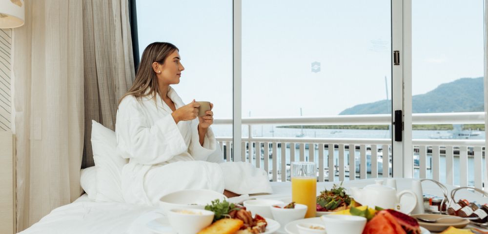 A woman in a robe sits on a bed with breakfast, holding a mug. She gazes out a window at a serene ocean view, creating a calm, relaxed atmosphere.