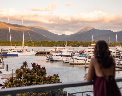 Woman in a red dress gazes at a serene marina with docked boats. Mountains and clouds glow in the sunset, creating a peaceful atmosphere.