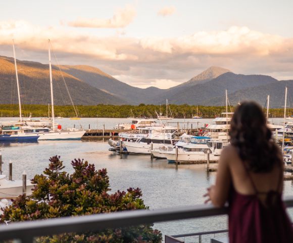 Woman in a red dress gazes at a serene marina with docked boats. Mountains and clouds glow in the sunset, creating a peaceful atmosphere.