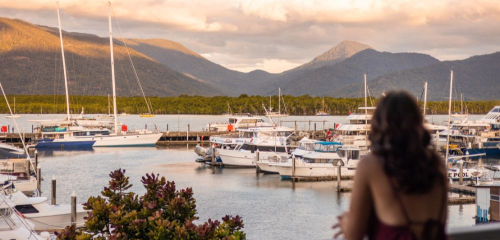 Woman in a red dress gazes at a serene marina with docked boats. Mountains and clouds glow in the sunset, creating a peaceful atmosphere.