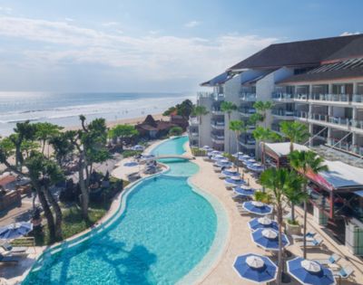 Aerial view of a beachfront resort with a large curving pool lined with blue umbrellas and lounge chairs. Ocean waves and lush greenery create a serene ambiance.