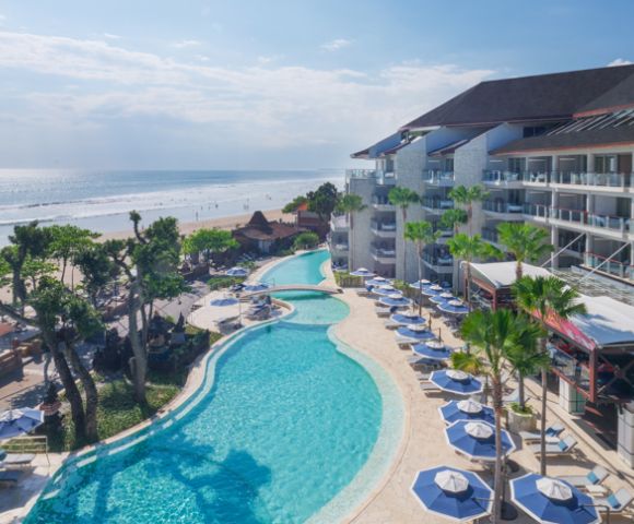 Aerial view of a beachfront resort with a large curving pool lined with blue umbrellas and lounge chairs. Ocean waves and lush greenery create a serene ambiance.