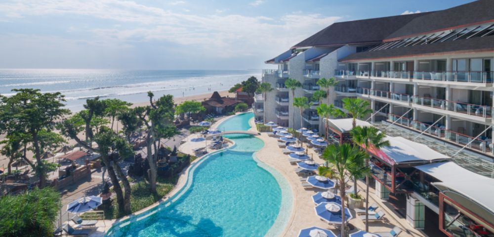 Aerial view of a beachfront resort with a large curving pool lined with blue umbrellas and lounge chairs. Ocean waves and lush greenery create a serene ambiance.