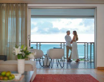 A couple stands on a balcony overlooking the ocean, embracing. The room has modern decor, with a vase of lilies and fruit bowl, evoking a serene, romantic ambiance.