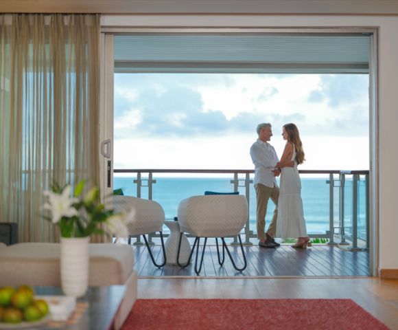 A couple stands on a balcony overlooking the ocean, embracing. The room has modern decor, with a vase of lilies and fruit bowl, evoking a serene, romantic ambiance.
