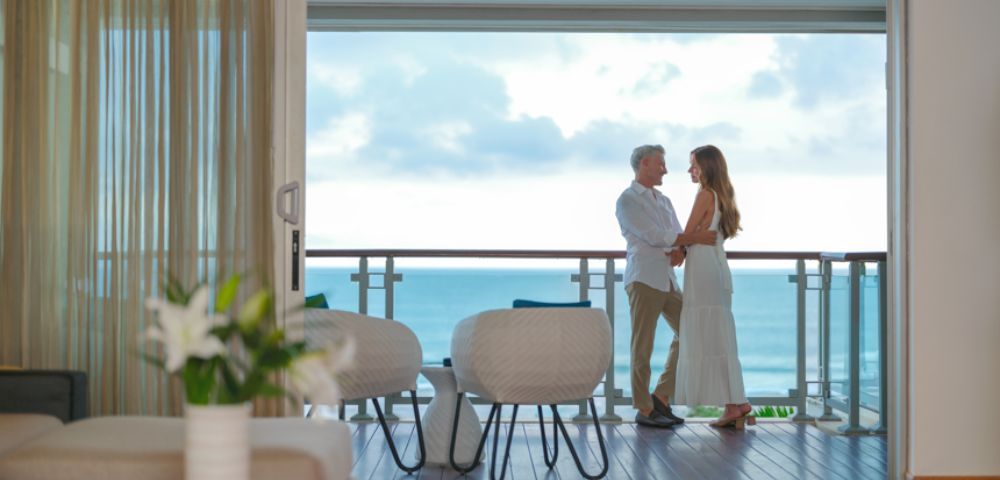 A couple stands on a balcony overlooking the ocean, embracing. The room has modern decor, with a vase of lilies and fruit bowl, evoking a serene, romantic ambiance.