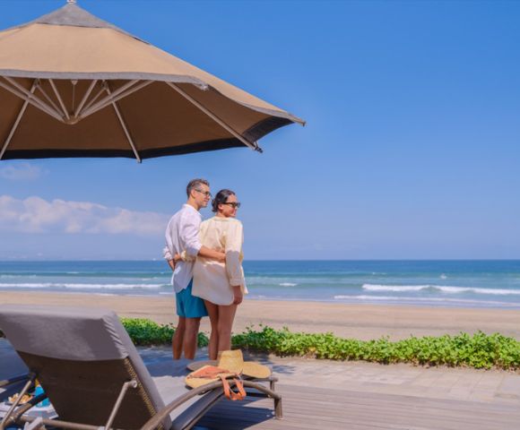 A couple in light summer clothing stands on a sunny beachfront deck, surrounded by lounge chairs and umbrellas, gazing at the tranquil ocean waves.