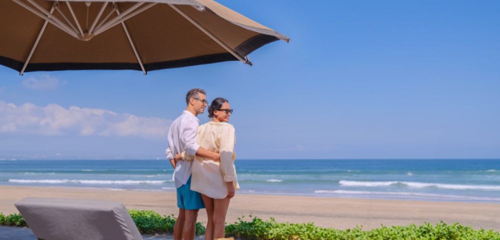 A couple in light summer clothing stands on a sunny beachfront deck, surrounded by lounge chairs and umbrellas, gazing at the tranquil ocean waves.