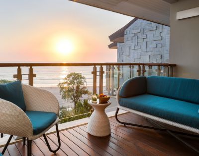 Cozy balcony view of a sunset over the ocean, with a teal-cushioned wicker chair and sofa. Warm wooden decking and serene, relaxing atmosphere.