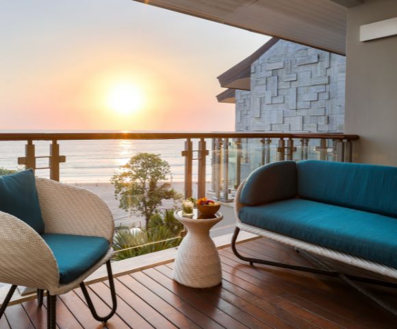 Cozy balcony view of a sunset over the ocean, with a teal-cushioned wicker chair and sofa. Warm wooden decking and serene, relaxing atmosphere.