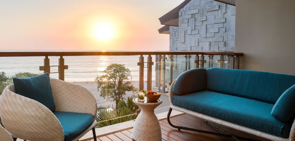 Cozy balcony view of a sunset over the ocean, with a teal-cushioned wicker chair and sofa. Warm wooden decking and serene, relaxing atmosphere.