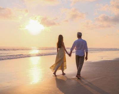 A couple walks hand in hand along a tranquil beach at sunset, casting long shadows on the sand. The sky is painted in soft pink and orange hues.