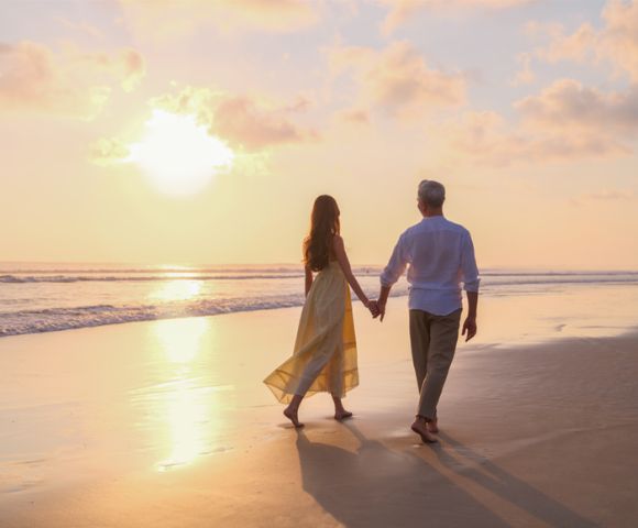 A couple walks hand in hand along a tranquil beach at sunset, casting long shadows on the sand. The sky is painted in soft pink and orange hues.