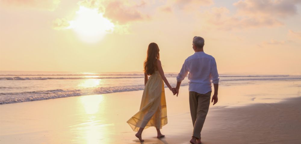 A couple walks hand in hand along a tranquil beach at sunset, casting long shadows on the sand. The sky is painted in soft pink and orange hues.