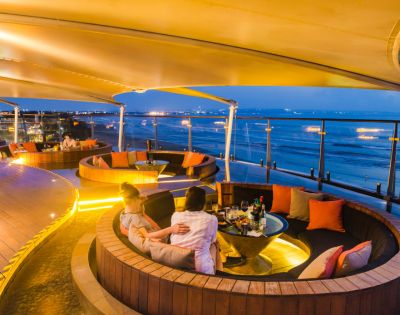 Cozy rooftop lounge at sunset with circular seating areas, warm yellow lighting, and people relaxing. Ocean view in the background enhances tranquility.