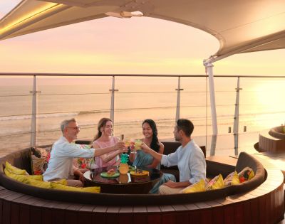 Four people sit in a round outdoor lounge with colorful pillows, raising glasses in a toast. They overlook a serene ocean at sunset, evoking joy and relaxation.