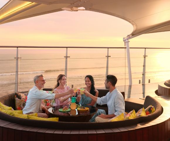 Four people sit in a round outdoor lounge with colorful pillows, raising glasses in a toast. They overlook a serene ocean at sunset, evoking joy and relaxation.