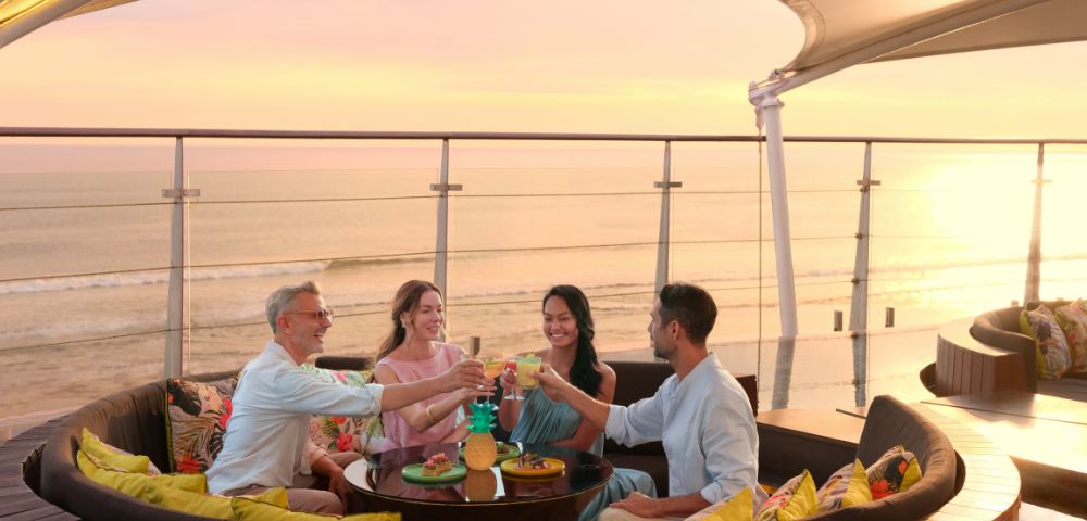Four people sit in a round outdoor lounge with colorful pillows, raising glasses in a toast. They overlook a serene ocean at sunset, evoking joy and relaxation.