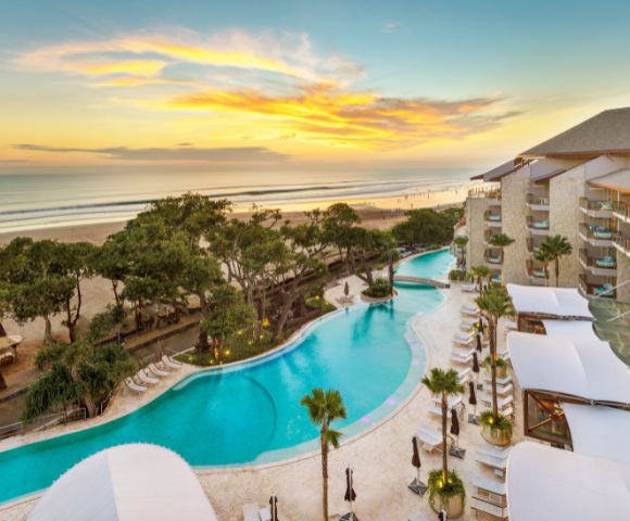 Aerial view of a luxurious beachfront resort at sunset. An expansive pool curves alongside palm trees and modern buildings, with a golden beach and calm ocean beyond.