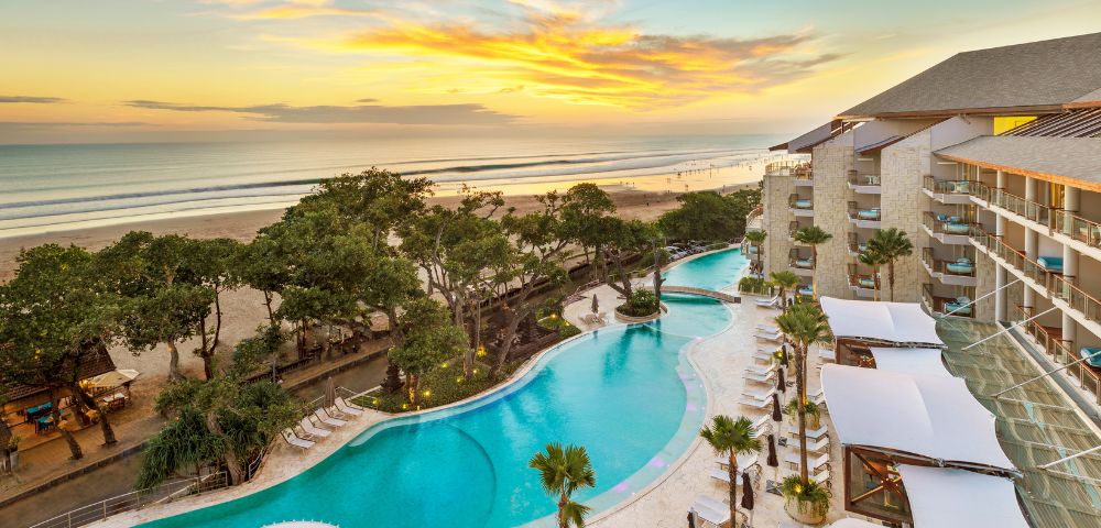 Aerial view of a luxurious beachfront resort at sunset. An expansive pool curves alongside palm trees and modern buildings, with a golden beach and calm ocean beyond.