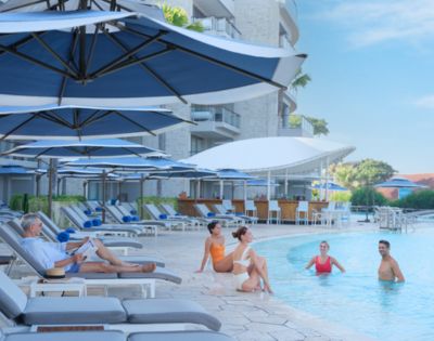 People enjoy a sunny day by a resort pool with loungers and large umbrellas. Some are seated poolside, others swim, surrounded by modern buildings.